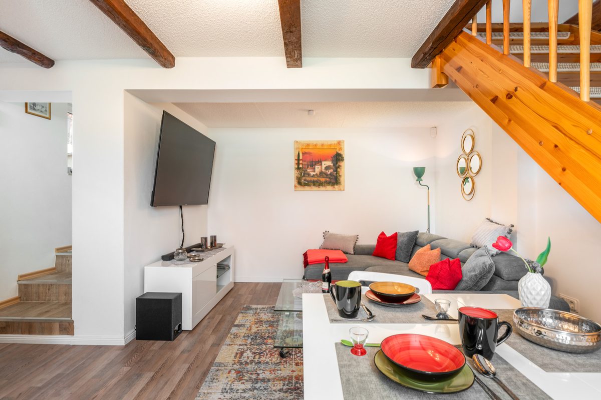 Heller Wohnbereich mit grauem Sofa, Fernseher, Essplatz und Holztreppe